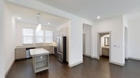 This two-story residence in The Colony offers a thoughtfully designed open-concept main level with clean, contemporary finishes. The kitchen is a central highlight, featuring light gray cabinetry extending to the upper walls, stainless steel appliances including a French-door refrigerator and range, and a mosaic tile backsplash that adds texture to the otherwise neutral palette. A kitchen island with built-in shelving provides additional storage and prep space, topped with a light-toned stone countertop. Pendant lighting hangs above the island, complementing the recessed ceiling lights throughout. Dark hardwood flooring runs seamlessly across the main living areas, creating a striking contrast against the white walls and light cabinetry. Natural light filters in through multiple windows fitted with white blinds. An arched doorway leads to an adjacent space that reveals a vanity area, suggesting a nearby bathroom with stone countertops and warm-toned cabinetry. The open layout connects the kitchen and living areas fluidly, lending the space a bright, airy ambiance with a modern yet welcoming character across both floors.