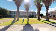 This single-floor space is situated within a two-story building set against a landscaped exterior featuring mature palm trees and a well-maintained lawn, evoking a warm, sun-drenched atmosphere. The building's façade presents a classic neutral-toned exterior with green-accented windows and open balcony railings, lending it a welcoming curb appeal.

Inside, the interiors are thoughtfully designed with a contemporary aesthetic. The dining area serves as a striking focal point, anchored by a richly veined green marble round table paired with woven rattan chairs atop a textured area rug. Polished concrete floors run throughout, adding an industrial yet refined quality to the space. Recessed lighting illuminates the crisp white walls, which are adorned with large-scale framed artwork that adds a curated, gallery-like sensibility.

A standout architectural feature is the sliding barn door with black hardware, offering a stylish and functional room divider that adds visual interest and character. Vintage-style double doors with ornate detailing connect to an adjacent room, where additional artwork and décor are visible, suggesting a cohesive and creatively appointed interior throughout. The overall ambiance is modern and eclectic, blending industrial materials with warm, artful touches.
