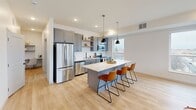 This thoughtfully designed single-floor residence offers a cohesive, contemporary aesthetic throughout. The open-concept layout connects the kitchen, dining, and living areas into one fluid, light-filled space. The kitchen is outfitted with gray shaker-style cabinetry, open shelving, stainless steel appliances, a white quartz countertop, and a central island with bar seating beneath industrial-style pendant lighting. Light hardwood flooring extends throughout the main living areas, complementing the clean white walls and modern finishes.

The dining area is a standout feature, framed by oversized windows on multiple sides that frame sweeping urban views and flood the space with natural light. A tray ceiling detail adds architectural interest overhead. The living room offers a relaxed, open feel with generous wall space and easy access to other areas of the unit.

Both bedrooms are carpeted for comfort and feature large windows with urban outlooks, providing brightness and a strong connection to the surrounding cityscape. Ample storage is available through a spacious walk-in closet with wire shelving systems, as well as an additional sliding-door closet. The full bathroom features a gray double vanity with quartz countertops, black hardware accents, pendant lighting, and a shower-tub combination.