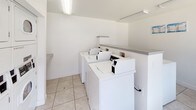 Located in Las Vegas, Nevada, this single-story space includes a functional laundry room designed with practicality in mind. The laundry area is outfitted with multiple top-loading washing machines positioned along one wall, providing ample capacity for laundry needs. The room features clean white walls and light-toned ceramic tile flooring, lending a bright and tidy atmosphere throughout. Overhead fluorescent lighting ensures the space is well-lit, contributing to a clean, utilitarian aesthetic. A built-in countertop or shelf runs along one side of the room, offering a convenient surface for folding or staging laundry. The overall layout is straightforward and efficient, maximizing usability within a compact footprint. The all-white color scheme of both the walls and appliances creates a cohesive, orderly appearance. This laundry space is well-suited for communal or shared use, reflecting a no-frills, functional design approach typical of shared residential or hospitality facilities in the Las Vegas area.