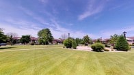 This single-level outdoor space presents a thoughtfully landscaped common area that combines functional design with natural beauty. The centerpiece of the grounds is a broad, well-kept lawn that stretches generously across the property, offering an open and inviting atmosphere under an expansive sky.

Mature trees are strategically positioned throughout the grounds, providing natural shade and a sense of established greenery. Neatly trimmed ornamental shrubs and carefully maintained garden beds add structure and visual interest to the landscape, reflecting consistent upkeep and attention to detail.

Paved walkways wind through the grounds, lined with classic black lamp posts that suggest safe and pleasant evening use. Wooden benches are placed along the paths, offering comfortable spots to rest and enjoy the surroundings. The overall layout encourages leisurely strolls and outdoor relaxation.

In the background, brick residential buildings with dark rooflines frame the space, contributing a warm, community-oriented character. The architecture is consistent and well-integrated into the landscape. The combination of open lawn, mature plantings, and thoughtfully placed amenities creates a serene, park-like environment that serves as an appealing communal outdoor retreat.