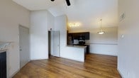This single-floor space makes a striking impression with its soaring vaulted ceilings that create an expansive, airy atmosphere throughout the open-concept layout. Rich, warm hardwood flooring flows seamlessly across the living and kitchen areas, anchoring the space with a sense of natural warmth and character.

The kitchen is a standout feature, appointed with dark espresso cabinetry that contrasts elegantly against light granite countertops. A peninsula with a built-in sink serves as both a functional workspace and a casual gathering point, complemented by a stylish pendant light overhead. Black appliances, including a built-in microwave and range, along with a stainless steel refrigerator, complete the well-equipped cooking space. A track lighting fixture above provides focused illumination across the kitchen.

The living area benefits from the dramatic ceiling height, enhanced further by a ceiling fan that aids in air circulation. Crisp white walls keep the space feeling bright and open, while a door off the main living area leads to an additional room or storage space. The overall ambiance is modern yet inviting, blending practical design with clean, contemporary finishes that create a comfortable and functional home environment.