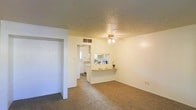 This single-floor residence offers a straightforward and functional layout designed for comfortable everyday living. The main living area is finished with wall-to-wall carpeting and neutral cream-toned walls, creating a bright and clean atmosphere. A ceiling fan with an integrated light fixture is centrally positioned, providing both air circulation and ambient lighting to the space.

A defining feature of the layout is the pass-through counter connecting the living area to the kitchen, allowing for an open and communicative flow between spaces. The kitchen, visible through both the pass-through and an adjacent doorway, is equipped with light wood cabinetry and receives natural light from a window, contributing to its airy feel. A small ledge along the pass-through provides a convenient surface for casual dining or serving.

The textured popcorn ceiling is consistent throughout, adding a classic residential character to the interior. A hallway visible from the living area suggests additional rooms beyond the main space. The overall ambiance is clean, neutral, and move-in ready, with a practical design that maximizes the use of the available floor plan on a single level.