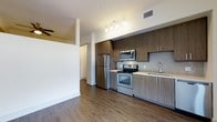 This single-floor unit combines functional design with modern finishes throughout its open layout. Dark hardwood-style flooring runs continuously across the space, providing a cohesive and polished look from room to room.

The kitchen is outfitted with stainless steel appliances, including a refrigerator, range, and microwave, complemented by medium-toned wood cabinetry with sleek hardware. A tile backsplash adds texture behind the range, while a single-basin sink and under-cabinet track lighting complete the functional workspace.

The living area features a vaulted ceiling section with a ceiling fan and light fixture, contributing to an airy, open feel. Natural light filters in through a window, brightening the neutral cream-colored walls. Multiple doorways off the main living space provide access to additional areas.

Storage is a notable highlight, with a spacious walk-in closet fitted with wire shelving, alongside additional louvered and panel-door closets. An in-unit laundry closet houses a stacked washer and dryer, adding everyday convenience. White panel doors with clean trim throughout maintain a consistent, contemporary aesthetic across the unit.