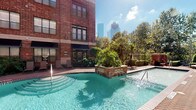 This amenity space is thoughtfully designed to blend industrial character with modern comfort. The outdoor pool area serves as a standout feature, set against the backdrop of a classic red brick building and framed by lush palm trees and greenery. A central brick fountain island with water features adds visual interest to the pool, while the surrounding deck provides space to relax. The urban skyline rises dramatically in the background, creating a striking contrast with the natural landscaping and wrought iron fencing that encloses the area.

Inside, the lounge area showcases exposed red brick accent walls paired with large, steel-framed windows that flood the space with natural light and offer views of the surrounding greenery. Comfortable gray upholstered sofas with geometric patterned throw pillows are arranged to encourage conversation and relaxation. The interior transitions seamlessly between zones, with one side featuring contemporary gray paneled walls adorned with a sculptural medallion, and track lighting overhead.

An adjacent business center offers a functional workspace, featuring built-in shelving lined with books set against an exposed brick wall, along with a desk and workstation. A wall-mounted flat-screen television near the entrance adds entertainment options to this well-rounded communal facility.