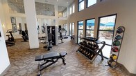This fitness center is a thoughtfully designed, single-floor facility that combines functionality with an open, welcoming atmosphere. The space is defined by its dramatically high ceilings, which lend an airy, expansive feel throughout. Large windows line the walls, flooding the interior with natural light and offering views of an outdoor patio area complete with seating and shade umbrellas — creating an energizing environment for workouts.

The main workout floor is finished with durable patterned carpeting and is divided into distinct functional zones. A dedicated cardio area houses multiple treadmills and a rowing machine, all positioned to take advantage of the natural light streaming in from the exterior windows. Nearby, a selection of stationary bikes and elliptical machines provide additional low-impact cardio options. Full-length mirrors are strategically placed throughout to visually expand the space and assist with form during exercise.

The strength training zone features a multi-station cable machine, an adjustable weight bench, a tiered dumbbell rack with a comprehensive range of free weights, and a medicine ball rack. Decorative framed artwork adorns the walls, adding a polished, upscale touch to the functional environment.

The facility also includes additional rooms accessible via interior doors, which appear to serve as support spaces. A flat-screen television is mounted for entertainment. The overall design balances a clean, contemporary aesthetic with practical, well-organized equipment placement.
