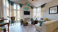 This single-level community lounge is thoughtfully designed to offer both comfort and functionality in an inviting, contemporary setting. The space is defined by its soaring vaulted ceilings in the main sitting area, which amplify the sense of openness and draw the eye upward toward decorative wall art composed of teal and gold medallions. Floor-to-ceiling windows dressed in striped teal, gold, and cream drapery flood the seating area with abundant natural light while framing views of the surrounding outdoor landscape. The sitting area features a generously sized neutral sectional sofa accented with teal and chartreuse throw pillows, complemented by a dark metal coffee table and eclectic accent chairs upholstered in bold geometric patterns. Light wood-look flooring extends throughout, tying all areas together seamlessly. A long bar-height counter with gray upholstered barstools serves as a central gathering point, anchored by pendant lighting overhead and a teal accent wall beyond. A compact refreshment alcove with granite countertops, warm wood cabinetry, and mosaic tile backsplash provides a convenient self-serve station. Framed artwork adorns the walls throughout, and the overall color palette of soft taupe, teal, and gold lends the lounge a polished, welcoming ambiance.