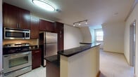 This single-floor residence offers a thoughtfully designed layout that balances functionality and comfort. The kitchen serves as a central highlight, showcasing rich dark mahogany-toned cabinetry that extends floor to ceiling, providing ample storage. Dark granite countertops contrast elegantly with the stainless steel appliances, including a full-size refrigerator, range, and dishwasher. A peninsula counter with a matching granite surface creates a natural division between the kitchen and the adjoining living area, which features neutral-toned carpeting that adds warmth underfoot. Track lighting and flush-mount ceiling fixtures illuminate the space throughout. The bedroom is generously proportioned with soft carpeting, a ceiling fan with integrated lighting, and a window with horizontal blinds that allows natural light to filter in. The angled roofline adds a distinctive architectural character to the room. The bathroom is well-equipped, featuring dark cabinetry with a clean white countertop and integrated sink, a large vanity mirror, and a full soaking tub with a tiled surround and shower fixture. Light tile flooring runs through the kitchen and bathroom, while plush carpet defines the living and sleeping areas, creating a cohesive and inviting overall ambiance.