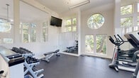 This fitness center in Columbia, Maryland, offers a thoughtfully designed workout environment that balances architectural appeal with functional exercise equipment. The space is defined by its striking double-height ceilings, which lend an open, airy quality throughout. A standout architectural feature is the large circular window positioned prominently on one wall, complemented by an array of tall rectangular windows that line multiple sides of the room, flooding the interior with natural light and framing views of the surrounding mature trees and greenery.

The flooring throughout is durable rubber gym matting in a dark charcoal tone, providing a practical and cushioned surface suitable for a range of exercise activities. The white walls and bright, clean aesthetic reinforce a fresh, modern atmosphere. Full-length mirrors are installed along one wall, enhancing the sense of space and supporting strength training.

Equipment includes a selection of Life Fitness elliptical trainers, upright and recumbent stationary bikes, and adjustable weight benches. A dedicated free weights area houses a tiered dumbbell rack with a comprehensive range of weights. Wall-mounted flat-screen televisions provide entertainment during workouts. Ceiling fans and linear light fixtures ensure comfort and adequate illumination. A staircase entry adds dimension to the layout, with the weight area partially visible from the upper landing.