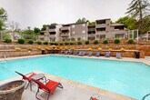 Located at 414 Point Return Dr in Ballwin, Missouri, this outdoor pool facility offers a well-appointed recreational retreat designed for relaxation and social gathering. The centerpiece is a generously sized swimming pool with clear, aqua-blue water, accessible via in-pool entry steps with handrails. The surrounding concrete deck provides ample space for lounging and entertaining, furnished with an array of blue reclining chaise lounges positioned along the pool's perimeter, as well as round patio tables paired with vibrant red and teal chairs, several topped with colorful umbrellas for shade. A pool house structure on one side provides utility access and additional amenities. The space is framed by a striking multi-tiered stone retaining wall, landscaped with neatly trimmed round shrubs, ornamental grasses, and seasonal plantings, which adds both visual interest and a sense of enclosure. Mature trees surround the area, offering natural shade and a lush green backdrop. A chain-link fence encloses the perimeter for safety. The overall setting is enhanced by views of the adjacent multi-story residential building, with balconies overlooking the pool area, creating a cohesive community atmosphere throughout.