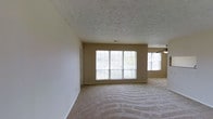 This single-floor space offers a clean, functional layout with a warm and inviting atmosphere. The main living area is generously proportioned, featuring neutral-toned wall-to-wall carpeting that adds comfort underfoot and complements the soft, beige-toned walls throughout. A textured popcorn ceiling adds a classic architectural detail overhead, while a ceiling fan provides practical air circulation and subtle visual interest in the corner of the room. Natural light is a standout feature of this space, with a large multi-pane picture window dominating the main wall and an additional side window allowing ample daylight to fill the room, creating a bright and airy feel. The windows also frame views of the surrounding greenery outside, connecting the interior to the natural environment. The open layout provides a versatile canvas, suitable for a variety of furniture arrangements and uses. Neutral finishes throughout — from the walls to the flooring — create a cohesive, move-in-ready aesthetic that is easy to personalize. Overall, the space conveys a sense of simplicity and practicality, well-suited for comfortable everyday living.