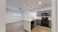 This thoughtfully designed one-bedroom, one-bathroom unit offers a smart and efficient layout across a single floor. The open-concept living and kitchen area serves as the heart of the space, anchored by a peninsula with a striking granite countertop that provides both additional workspace and casual seating. The kitchen is outfitted with gray shaker-style cabinetry, a full suite of black appliances including a range, microwave, dishwasher, and refrigerator, all complemented by a clean white backsplash. Three stylish pendant lights hang above the peninsula, adding a modern touch, while recessed lighting and a ceiling fan with a light kit illuminate the adjoining living area. A window with blinds brings in natural light to brighten the open space. Warm wood-look flooring flows consistently throughout the unit, tying each room together seamlessly. The bedroom is a comfortable, well-proportioned retreat with its own ceiling fan, a bright window, and direct access to both a dedicated closet and the hallway. The bathroom features a gray vanity with a granite-topped sink, large-format tile surrounding a full soaking tub with shower, and matching wood-look flooring for a cohesive, clean aesthetic. A separate closet and hallway provide practical storage and circulation throughout the unit.