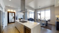 This contemporary single-floor space exemplifies modern urban living through its open-concept layout and refined design aesthetic. The kitchen serves as a striking centerpiece, anchored by a generous waterfall-edge island finished in pristine white stone and equipped with a built-in gas cooktop. A stainless steel island range hood rises dramatically toward the high ceilings, reinforcing the kitchen's professional-grade character. White cabinetry with sleek hardware completes the clean, minimalist look, while warm hardwood flooring flows seamlessly throughout, providing a natural contrast to the cool, contemporary palette. Pendant lighting adds refined illumination across the kitchen and adjacent areas. The open floor plan connects the kitchen effortlessly to a dedicated dining area, where a long rectangular table is surrounded by modern acrylic ghost chairs, and further into a comfortable living room furnished with a classic dark sectional sofa. Floor-to-ceiling windows wrap the space, flooding every corner with abundant natural light and offering sweeping panoramic city views. Neutral roller shades provide flexible light control without compromising the sense of openness. The overall ambiance is polished and sophisticated, blending high-end finishes with an airy, light-filled atmosphere ideal for both daily living and entertaining.