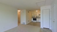 This single-floor unit offers an open and functional layout, seamlessly connecting the living area to the kitchen. The living space features wall-to-wall carpet in a neutral beige tone, contributing to a warm and understated aesthetic. A floor vent and clean white walls give the space a simple, practical character.

The kitchen is distinguished by tile flooring, providing a clear visual and functional separation from the carpeted living area. It is equipped with white cabinetry arranged along the upper and lower walls, offering ample storage. Standard appliances include a white electric range and a built-in microwave above it, as well as a full-size refrigerator positioned at the edge of the space. A single-basin sink sits beneath upper cabinets along the adjacent wall, completing a compact yet efficient kitchen configuration.

A hallway visible from the main living area leads toward additional rooms, with a door visible at its end, suggesting private spaces such as a bedroom or bathroom beyond. Ceiling-mounted light fixtures provide consistent overhead illumination throughout. The overall ambiance is clean, straightforward, and move-in ready.