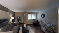 This single-floor space centers around a well-appointed living room that combines comfort and style. The room features rich, dark hardwood flooring that contrasts nicely with the neutral beige and gray walls, creating a warm and inviting atmosphere. A textured popcorn ceiling adds a classic touch overhead.

Natural light filters into the space through a centrally positioned window dressed with white horizontal blinds and dark wood trim, lending the room a grounded, defined look. A wall-mounted air conditioning unit provides climate control convenience.

The living area is furnished with a substantial gray sectional sofa, a sleek black coffee table atop a light area rug, and a black accent chair with a decorative cushion. A patterned accent chair in the corner adds visual interest. Decorative touches include a sunburst-style mirror on the wall, a black shelving unit, and carefully placed accent pieces that enhance the overall aesthetic.

A floor lamp in the corner provides supplemental lighting, contributing to the room's warm ambiance. The space balances functional living with thoughtful design details throughout.
