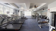 This single-floor fitness center is thoughtfully designed to support a comprehensive workout experience across several distinct functional zones. The main cardio area features a row of Star Trac treadmills and elliptical machines positioned along a wall of windows, allowing natural light to fill the space and providing views of lush greenery outside. Ceiling fans mounted within decorative tray ceilings with crown molding add an architectural elegance uncommon in typical gym settings, while recessed lighting ensures consistent illumination throughout.

The strength training section is equipped with a variety of cable and resistance machines, including multi-functional units and dedicated benches, all arranged to allow ample space for safe movement between stations. Floor-to-ceiling mirrors line one wall, visually expanding the space and enabling users to monitor their form during exercises. A dumbbell rack further rounds out the free weights area.

A separate room provides a dedicated space for group fitness or stretching activities, complete with step platforms, resistance accessories, a wall-mounted television, and a large window that brings in natural light. The consistent blue-toned carpet flooring and soft gray walls create a calm, focused atmosphere throughout the entire facility, making it a functional and inviting environment for a wide range of fitness activities.