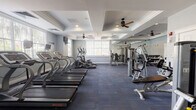 This single-floor fitness center is thoughtfully designed to support a comprehensive workout experience across several distinct functional zones. The main cardio area features a row of Star Trac treadmills and elliptical machines positioned along a wall of windows, allowing natural light to fill the space and providing views of lush greenery outside. Ceiling fans mounted within decorative tray ceilings with crown molding add an architectural elegance uncommon in typical gym settings, while recessed lighting ensures consistent illumination throughout.

The strength training section is equipped with a variety of cable and resistance machines, including multi-functional units and dedicated benches, all arranged to allow ample space for safe movement between stations. Floor-to-ceiling mirrors line one wall, visually expanding the space and enabling users to monitor their form during exercises. A dumbbell rack further rounds out the free weights area.

A separate room provides a dedicated space for group fitness or stretching activities, complete with step platforms, resistance accessories, a wall-mounted television, and a large window that brings in natural light. The consistent blue-toned carpet flooring and soft gray walls create a calm, focused atmosphere throughout the entire facility, making it a functional and inviting environment for a wide range of fitness activities.