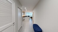 This single-floor high-rise residence offers a well-organized layout that begins at a welcoming entryway hallway finished with large-format white ceramic tile. A navy upholstered bench near the entry provides a practical landing spot, while recessed lighting illuminates the path toward the main living areas.

The kitchen is thoughtfully designed with white cabinetry, dark granite countertops, and a full suite of stainless steel appliances, including a refrigerator, range, built-in microwave, and dishwasher. Pendant lighting accents the space, which transitions seamlessly toward the open living and dining area.

The living area is a standout feature, anchored by dramatic floor-to-ceiling curved glass windows that frame expansive, elevated city views. Wood-look flooring, a compact dining set, and leather seating arrangements make this an inviting and visually striking space filled with abundant natural light.

The primary bedroom continues the curved glass window theme, offering direct access to a balcony and soft carpeted flooring. A second bedroom provides a quieter, carpeted retreat with ample closet access. The bathroom features tile flooring, a dark granite vanity, and a glass-enclosed walk-in shower.