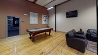 This single-floor entertainment space is divided into two distinct yet complementary areas: a spacious game room and a dedicated private theater. The game room is anchored by a full-size pool table set atop warm hardwood flooring, surrounded by soaring ceilings with exposed structural columns and industrial-style ductwork that lend the space a loft-like character. Large floor-to-ceiling glass partitions allow natural light to filter in, while a mounted flat-screen television, foosball table, bar-height seating, and a comfortable leather lounge area round out the amenities. Vintage decorative posters and artwork add personality throughout.

The theater room transitions into a more intimate atmosphere, featuring plush leather recliner seating arranged in rows with individual side tables for each guest. Exposed brick walls serve as a striking backdrop for a curated collection of classic movie posters, including titles such as E.T., Harry Potter, and Goodfellas. Recessed lighting and carpeted flooring enhance the cinematic ambiance, while a large display screen positioned at the front of the room completes the private screening experience. Together, the two areas create a well-appointed entertainment destination ideal for gatherings.
