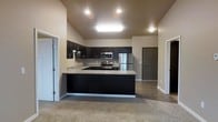 This single-floor space centers around a well-appointed kitchen that serves as the focal point of the open-concept layout. The kitchen features rich, dark cabinetry that extends to the upper walls, providing ample storage throughout. Light-toned countertops offer a striking contrast against the dark lower cabinets, creating a visually balanced aesthetic. Stainless steel appliances, including a refrigerator, range, and built-in microwave, are seamlessly integrated into the design.

The kitchen is arranged in a U-shaped configuration with a peninsula that extends outward, defining the boundary between the kitchen and the adjoining living area. Recessed lighting and a central skylight fixture illuminate the kitchen workspace effectively. The vaulted ceiling adds a sense of vertical openness, enhancing the overall spaciousness of the area.

The adjoining living space is finished with neutral-toned carpet, providing a warm transition from the kitchen's hard flooring. The open layout allows for flexible furniture arrangement. Multiple doorways visible from the main area suggest additional rooms, contributing to a functional and connected floor plan throughout the space.