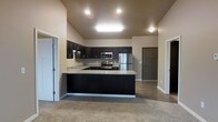 This single-floor space centers around a well-appointed kitchen that serves as the focal point of the open-concept layout. The kitchen features rich, dark cabinetry that extends to the upper walls, providing ample storage throughout. Light-toned countertops offer a striking contrast against the dark lower cabinets, creating a visually balanced aesthetic. Stainless steel appliances, including a refrigerator, range, and built-in microwave, are seamlessly integrated into the design.

The kitchen is arranged in a U-shaped configuration with a peninsula that extends outward, defining the boundary between the kitchen and the adjoining living area. Recessed lighting and a central skylight fixture illuminate the kitchen workspace effectively. The vaulted ceiling adds a sense of vertical openness, enhancing the overall spaciousness of the area.

The adjoining living space is finished with neutral-toned carpet, providing a warm transition from the kitchen's hard flooring. The open layout allows for flexible furniture arrangement. Multiple doorways visible from the main area suggest additional rooms, contributing to a functional and connected floor plan throughout the space.