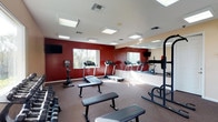 This fitness center is designed to support a full range of workout routines in a focused and functional environment. The space is defined by bold, deep-red accent walls that energize the room, complemented by neutral carpeted flooring that provides comfort underfoot during exercise. A bank of large windows lines one wall, inviting an abundance of natural light and offering views of the surrounding greenery, creating an uplifting atmosphere for workouts. Full-length mirrors are mounted along an adjacent wall, enhancing the sense of space and allowing users to monitor their form during training. The cardio section features multiple treadmills arranged to take advantage of the natural light from the windows. The strength training area is well-appointed with a set of dumbbells racked along one wall, flat weight benches, and a multi-function power rack, catering to a variety of resistance training needs. Overhead, recessed flat-panel light fixtures ensure consistent, even illumination throughout the space. A wall-mounted flat-screen television adds a convenient entertainment option during workouts. The overall ambiance is motivating and practical, making this fitness center a comprehensive facility for both cardio and strength training.