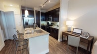 This single-floor space showcases a thoughtfully designed open-concept layout that seamlessly integrates the kitchen and living areas. The kitchen is a standout feature, anchored by a central island with a granite countertop, an undermount stainless steel sink, and bar seating — ideal for casual dining or entertaining. Dark espresso cabinetry extends to the ceiling, providing generous storage, while a light tile backsplash creates a striking contrast. Stainless steel appliances, including a French door refrigerator with an ice and water dispenser, a gas range, and an over-the-range microwave, complete the kitchen's modern aesthetic. Track lighting overhead ensures the workspace is well-lit, complemented by a pendant light above the island. Warm hardwood-style flooring runs throughout, adding continuity and a welcoming feel to the space. Adjacent to the kitchen, a built-in desk area with a wood surface and task lamp provides a dedicated workspace, making the unit as functional as it is stylish. Clean white walls and neutral tones contribute to a bright, open ambiance throughout.