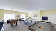 This single-floor recreational space is thoughtfully designed to accommodate a variety of games and social activities. The room is generously proportioned, offering ample space for multiple games to be enjoyed simultaneously without feeling crowded. The interior features light yellow walls accented by crisp white crown molding and a smooth white ceiling with recessed lighting, creating a bright and welcoming atmosphere. Carpeted flooring adds comfort underfoot throughout the space. Large windows along one wall allow natural light to fill the room, enhancing its open and airy feel. The space is outfitted with a full-size billiards table, a regulation ping-pong table, and a foosball table, catering to a wide range of recreational interests. Additional seating and a small table are available for casual conversation or spectating. A chalkboard mounted on the wall serves as a practical scorekeeping surface, while framed artwork adds a decorative touch to the walls. A dark wood desk or console unit is positioned near the windows, offering additional functionality. Overall, this game room provides a versatile and inviting environment well-suited for leisure, entertainment, and community engagement.