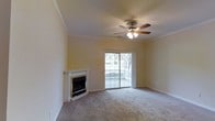 This single-story space centers around a well-appointed living area that combines comfort and functionality. The room is finished with wall-to-wall carpeting in a neutral tone, complemented by soft yellow walls and crisp white crown molding that adds a refined architectural detail along the ceiling perimeter. A textured ceiling overhead contributes subtle visual interest throughout the space.

A corner fireplace with a clean white surround serves as a focal point, offering both aesthetic appeal and practical warmth. Above, a ceiling fan with a built-in light fixture provides year-round comfort and ambient lighting. Natural light enters generously through a sliding glass door, which opens to an outdoor deck or balcony, framing views of surrounding trees and the exterior landscape beyond. The sliding door not only brightens the interior but also creates a seamless visual connection between the indoor living space and the outdoors.

The overall ambiance is calm and inviting, with a cohesive palette of neutral tones throughout. The combination of carpeted flooring, crown molding, fireplace, and outdoor access makes this a versatile and comfortable living environment.