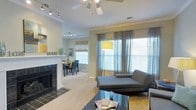 This single-floor residence offers a well-designed, open-concept layout that seamlessly connects the living and dining areas, creating a warm and inviting atmosphere throughout. The living room serves as the heart of the home, anchored by a classic white-mantled fireplace with a striking black tile surround, adding both character and comfort to the space. Plush carpeting underfoot enhances the cozy ambiance, while a ceiling fan keeps the area comfortable year-round. Large windows dressed in sheer grey draping curtains allow generous amounts of natural light to fill the room, complementing the neutral wall tones. A bold yellow floor lamp adds a cheerful pop of color and modern flair to the living area. The dining space, visible through an open passage, features an intimate table setting with coordinating yellow accents, surrounded by dark dining chairs. Bamboo shades on the dining room windows add a natural, textured touch. Track lighting in both areas provides flexible, functional illumination. Crown molding and clean architectural details throughout lend a polished, finished look to the overall design, making this a comfortable and stylish single-level home.