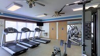 This fitness center offers a practical and well-organized space for exercise and wellness. The room is decorated with a distinctive striped accent wall in blue and burgundy, adding a touch of character to the functional environment. Ceiling fans are installed throughout, promoting air circulation and comfort during workouts.

The cardio area is outfitted with multiple treadmills lined along the windowed wall, allowing for natural light to brighten the space. A wall-mounted television provides entertainment for users during their sessions. A built-in water fountain is conveniently located near the center of the room for easy access during exercise.

A large wall mirror spans one side of the room, visually expanding the space and allowing users to monitor their form while using the strength and resistance equipment positioned nearby. The flooring is a durable, low-pile carpet suited for fitness use.

Two clearly marked, ADA-accessible restrooms are accessible directly from the fitness room, adding convenience and inclusivity to the facility. Overhead fluorescent lighting supplements the natural light from the windows, ensuring the space remains bright throughout the day. Overall, the fitness center is thoughtfully designed to be both functional and welcoming for a wide range of users.