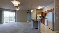 This single-floor residence showcases an open-concept design that seamlessly connects the living area and kitchen into one cohesive space. The living area is finished with wall-to-wall carpeting and features a ceiling fan with an overhead light fixture, contributing to a comfortable, functional ambiance. Natural light filters in through a sliding glass door that leads to an outdoor space, brightening the interior and providing a sense of openness.

The kitchen is well-appointed with warm medium-toned wood cabinetry lining the walls, offering ample storage. White appliances, including a range and an over-the-range microwave, are neatly integrated into the layout. A peninsula island with a dark countertop serves as the centerpiece of the kitchen, providing additional prep space and acting as a natural divider between the kitchen and living zones. Recessed and pendant lighting above the island adds focused illumination to the workspace.

A hallway visible from the main living space leads to additional rooms, with a paneled door providing access to further areas of the unit. The neutral gray wall tones throughout create a clean, understated backdrop that complements the overall layout.