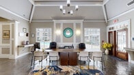 Located at 2742 Jeanetta Rd in Houston, TX, this single-floor community space offers a refined and welcoming environment defined by sophisticated architectural detailing and thoughtful design. The space is organized around a dining room and a living room, each contributing to a cohesive, upscale atmosphere.

The dining area serves as a functional centerpiece, anchored by a rich walnut-toned conference table surrounded by a mix of executive and guest chairs. A vibrant abstract area rug in navy, gold, and white adds a dynamic visual element underfoot. Paneled wainscoting with wall sconces and a striking teal circular mirror lend the room a polished, curated character, while large windows fitted with white blinds frame views of an outdoor pool area.

The living room is equally impressive, boasting soaring vaulted ceilings with a coffered tray detail and an elegant multi-light chandelier. Stone-tiled flooring flows throughout, complemented by neutral warm-gray walls and crisp white trim. Transom-topped doorways and French-style entry doors with sidelights flood the interior with natural light, while a wall-mounted television and a granite-topped counter add practical amenities. Comfortable seating vignettes with modern gray chairs create inviting conversation areas. Together, the rooms project a professional yet welcoming ambiance ideal for gatherings and collaboration.