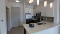 This single-floor unit showcases a well-designed open-concept kitchen that serves as the centerpiece of the living space. The kitchen is outfitted with a full suite of stainless steel appliances, including a French door refrigerator, an electric range, and an over-the-range microwave, all set against a backdrop of sleek, gray geometric tile backsplash. Crisp white cabinetry extends to the upper walls, providing generous storage space, while the lower cabinets offer a dark contrast that adds visual depth to the design. A spacious kitchen island with a light-toned countertop provides additional prep and seating space, and is equipped with a stainless steel undermount sink and a modern arc faucet. Pendant lighting and flush-mount ceiling fixtures illuminate the space effectively. The entryway, visible from the kitchen, features a solid panel door with contemporary hardware. Light wood-tone flooring runs throughout, complementing the neutral color palette of white and gray tones that give the space a clean, modern aesthetic.