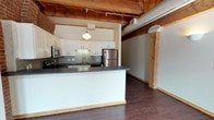 This two-floor loft-style space masterfully balances industrial character with contemporary finishes. Throughout the main level, rich hardwood flooring anchors an open layout, while exposed heavy timber ceiling beams and visible ductwork celebrate the building's historic industrial roots. The open-concept kitchen serves as a central hub, featuring white shaker-style cabinetry, dark granite countertops, a tile backsplash, and a full suite of stainless steel appliances including a range, built-in microwave, and refrigerator. A peninsula counter creates a natural divide between the kitchen and the expansive living area, which benefits from generous natural light filtering through the windows. Exposed red brick walls appear prominently across multiple areas, lending warmth and texture to the interior. The bathroom is finished with white tile flooring, a tub-shower combination, and a large vanity mirror. Upper-level spaces showcase the same exposed brick and timber beam aesthetic, with carpeted flooring offering a softer contrast. High clerestory windows on the upper level draw in additional natural light and reinforce the soaring sense of vertical space. The result is a distinctive space that honors its architectural heritage while providing modern, functional living areas.