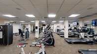 This comprehensive fitness center offers a generously sized, single-floor layout designed to accommodate a full range of workout activities. The main exercise area features a drop-panel ceiling with recessed lighting that evenly illuminates the space, supported by structural columns that define distinct workout zones throughout the floor. Durable, textured carpet covers much of the main floor, providing a practical surface for the extensive array of cardio and strength equipment. Rows of Spirit-brand treadmills and elliptical trainers are neatly arranged alongside stationary bikes, offering ample cardio options. Strength training is well represented with multi-station cable machines, weight benches, and a full dumbbell rack with free weights and kettlebells set on rubberized flooring. Wall-mounted flat-screen televisions are positioned throughout for entertainment. Framed artwork and potted plants add a welcoming touch to the otherwise functional environment. A separate studio room, enclosed by interior windows overlooking the main floor, features smooth hardwood flooring, full-length mirrored walls, and exercise mats, making it ideal for stretching, yoga, or group classes. The studio connects via a wood-trimmed glass door, completing a well-rounded and thoughtfully organized fitness facility.