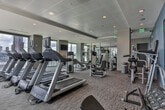 Located in the heart of Midtown Atlanta, this well-appointed fitness center offers a comprehensive workout environment across a single, thoughtfully organized floor. The facility is divided into distinct exercise zones, each designed to support a full range of training styles. A standout feature is the floor-to-ceiling glass walls that flood the space with natural light and frame sweeping panoramic views of the Atlanta skyline and surrounding cityscape, creating an inspiring backdrop for any workout.

The cardio area is equipped with treadmills and other aerobic machines positioned to take full advantage of the city views. A dedicated free weights section includes a complete dumbbell rack and adjustable benches set on protective rubber flooring, accompanied by a wall-mounted flat-screen television. The strength training zone features Precor cable and resistance machines, arranged to maximize usable floor space. Colorful kettlebells, stability balls, and additional functional training equipment are housed behind floor-to-ceiling glass panel walls, visible from the facility's entrance corridor.

A water station is conveniently located near the entrance for easy access. The interior design incorporates bold teal accent walls, neutral carpeted flooring, and recessed lighting throughout, contributing to a clean, modern ambiance. Mirrors are strategically placed along the walls to enhance the sense of space and support proper form during exercise.