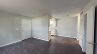 This single-floor residence offers a practical and well-organized layout designed for comfortable everyday living. The open living area greets you upon entry, featuring dark hardwood-style flooring and neutral gray walls that create a clean, versatile backdrop. The space flows seamlessly toward a pass-through kitchen area, where white cabinetry and a sink are visible, providing a functional connection between the living and cooking spaces.

The galley-style kitchen is efficiently designed with white cabinetry lining both sides, ample counter space, a double basin sink, and a white electric range with overhead ventilation. A window at the end of the kitchen allows for natural light and ventilation.

The bedrooms are carpeted in a neutral gray tone, offering a softer underfoot feel. One bedroom features a window that brings in natural light and views of surrounding greenery, while another is spacious with multiple outlets and flexible wall space. The primary bedroom area includes both a louvered closet door and an additional open closet, offering generous storage, with a hallway leading directly to the bathroom.

The bathroom is finished in white, featuring a single vanity with under-cabinet storage, a wall mirror with overhead lighting, and a bathtub. Throughout the unit, consistent neutral tones, white trim, and dark flooring create a cohesive and understated aesthetic.