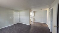 This single-floor residence offers a practical and well-organized layout designed for comfortable everyday living. The open living area greets you upon entry, featuring dark hardwood-style flooring and neutral gray walls that create a clean, versatile backdrop. The space flows seamlessly toward a pass-through kitchen area, where white cabinetry and a sink are visible, providing a functional connection between the living and cooking spaces.

The galley-style kitchen is efficiently designed with white cabinetry lining both sides, ample counter space, a double basin sink, and a white electric range with overhead ventilation. A window at the end of the kitchen allows for natural light and ventilation.

The bedrooms are carpeted in a neutral gray tone, offering a softer underfoot feel. One bedroom features a window that brings in natural light and views of surrounding greenery, while another is spacious with multiple outlets and flexible wall space. The primary bedroom area includes both a louvered closet door and an additional open closet, offering generous storage, with a hallway leading directly to the bathroom.

The bathroom is finished in white, featuring a single vanity with under-cabinet storage, a wall mirror with overhead lighting, and a bathtub. Throughout the unit, consistent neutral tones, white trim, and dark flooring create a cohesive and understated aesthetic.