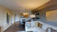 This single-floor residence showcases an open-concept layout that seamlessly connects the living, dining, and kitchen areas into one cohesive, functional space. The living area is anchored by a contemporary white sectional sofa accented with silver and gold throw pillows, paired with a tall floor lamp that provides warm ambient lighting. A built-in wall-mounted shelving unit in a deep gray tone adds a stylish focal point, offering display and storage space for decorative items and barware.

Adjacent to the living area, the dining space features a round dark table surrounded by modern upholstered chairs, illuminated overhead by an elegant chandelier with a classic pendant design. The kitchen, partially open to the dining area via a breakfast bar, is equipped with wood-toned cabinetry and stainless steel appliances, including a refrigerator, creating a practical and accessible cooking space.

Dark hardwood flooring runs throughout, providing a rich contrast to the light gray and white walls. The entry area is visible in the background, featuring a clean white door. The overall ambiance is modern, warm, and well-organized.