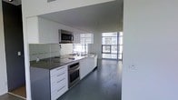 This contemporary two-floor space exemplifies modern urban living through its clean lines, minimalist aesthetic, and high-end finishes throughout. The open-concept kitchen and living area serves as the heart of the main floor, where glossy white cabinetry and stainless steel appliances — including a built-in oven, cooktop, microwave, and dishwasher — are seamlessly integrated against sleek dark countertops. The kitchen flows naturally into the living area, creating a cohesive and functional space ideal for both everyday living and entertaining. Large floor-to-ceiling windows bathe the interior in abundant natural light while offering expansive views of the surrounding cityscape, enhancing the sense of openness. The dark-toned flooring provides a striking contrast against the crisp white walls and cabinetry, adding depth and sophistication to the overall design. Recessed lighting complements the natural light, ensuring the space remains bright and welcoming at all hours. The two-floor layout provides a clear sense of vertical space, contributing to an airy and elevated atmosphere throughout. Every element of this space has been carefully considered to deliver a refined, modern environment with a distinctly urban character.