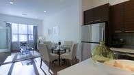 This single-floor residence showcases a thoughtfully designed open-concept layout that integrates the kitchen, dining, and living areas into one cohesive living space. The kitchen is equipped with stainless steel appliances, including a full-sized refrigerator topped with dark-finish upper cabinetry, providing ample storage. A white countertop anchors the kitchen area, offering functional workspace that flows naturally into the dining zone.

The dining area features a round glass-top table surrounded by upholstered cream chairs with nailhead trim detailing, lending an elegant yet approachable aesthetic. Hardwood flooring runs throughout, adding warmth and visual continuity across the space.

Natural light is a defining characteristic, streaming in through large windows dressed with grey curtains in the living area beyond. The crisp white walls are accented with framed artwork, enhancing the clean, modern atmosphere. Recessed lighting supplements the natural light, ensuring the space remains bright and well-illuminated at all times. The overall ambiance is contemporary and inviting, with a neutral color palette that creates a versatile backdrop for a variety of interior styles.