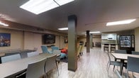 This single-floor space is designed as an open-plan communal area that seamlessly integrates multiple functional zones within one continuous layout. The interior is defined by bold dark structural columns that rise to a flat ceiling fitted with recessed and fluorescent panel lighting, providing consistent, even illumination throughout the space.

The flooring transitions between light wood-look planks across the main areas and a patterned area rug beneath a casual lounge arrangement, adding visual variety and zoning without physical barriers. The lounge area features upholstered seating in teal and neutral tones grouped around a wall-mounted flat-screen television, creating a relaxed gathering spot.

Adjacent to the lounge, a dining area with a round table and contemporary high-back chairs offers a designated space for meals or meetings. Further into the space, a kitchenette with white cabinetry, a countertop, and bar-height seating provides a practical food preparation and casual dining station. Additional seating clusters near the rear suggest flexible use for a variety of group activities. The neutral color palette of warm tans and grays contributes to a calm, functional ambiance suited to shared or communal use.