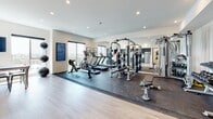 This well-appointed fitness facility offers a thoughtfully designed, single-floor layout that combines functionality with a clean, modern aesthetic. The centerpiece of the space is a generously sized gym area, featuring rubber flooring over light hardwood and an impressive array of equipment — including a multi-station cable and weight machine, treadmills, an elliptical, a rowing machine, and a full dumbbell rack. Floor-to-ceiling mirrors line one wall, amplifying the sense of space and allowing for form monitoring during workouts. Large windows invite natural light and offer sweeping panoramic views, creating an energizing atmosphere. Recessed lighting throughout ensures the space remains bright and functional at any hour. Adjacent to the gym floor is a sleek kitchenette outfitted with dark wood-grain cabinetry, stainless steel appliances including a refrigerator, gas range, and microwave, and a light marble-style countertop paired with a white geometric tile backsplash. Light hardwood flooring flows seamlessly through this area, maintaining the cohesive contemporary design. A dedicated restroom rounds out the facility, finished with the same light hardwood flooring, clean white walls, accessibility grab bars, and tasteful black-and-white artwork for a polished, curated feel throughout.