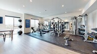This well-appointed fitness facility offers a thoughtfully designed, single-floor layout that combines functionality with a clean, modern aesthetic. The centerpiece of the space is a generously sized gym area, featuring rubber flooring over light hardwood and an impressive array of equipment — including a multi-station cable and weight machine, treadmills, an elliptical, a rowing machine, and a full dumbbell rack. Floor-to-ceiling mirrors line one wall, amplifying the sense of space and allowing for form monitoring during workouts. Large windows invite natural light and offer sweeping panoramic views, creating an energizing atmosphere. Recessed lighting throughout ensures the space remains bright and functional at any hour. Adjacent to the gym floor is a sleek kitchenette outfitted with dark wood-grain cabinetry, stainless steel appliances including a refrigerator, gas range, and microwave, and a light marble-style countertop paired with a white geometric tile backsplash. Light hardwood flooring flows seamlessly through this area, maintaining the cohesive contemporary design. A dedicated restroom rounds out the facility, finished with the same light hardwood flooring, clean white walls, accessibility grab bars, and tasteful black-and-white artwork for a polished, curated feel throughout.