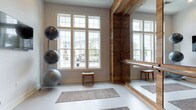 This single-floor fitness and yoga studio is thoughtfully designed to blend rustic warmth with clean, modern functionality. The space is anchored by a striking architectural feature: a large reclaimed wood-framed mirror wall fitted with metal ballet barres, providing both a visual focal point and a practical tool for barre and dance-inspired workouts. The contrast between the rich, dark wood tones and the crisp white walls and polished light floors creates a welcoming, balanced ambiance throughout.

Large multi-pane windows line the walls, flooding the studio with abundant natural light and offering views of the surrounding neighborhood, contributing to an open, airy atmosphere. The smooth, light-toned flooring is punctuated by a series of woven, textured mats arranged to accommodate multiple participants simultaneously.

Equipment is neatly organized throughout the space, including a rack of stability balls in varying sizes, foam rollers stored in a wire basket, and small wooden benches that serve both functional and decorative purposes. A wall-mounted flat-screen television is positioned for guided workout viewing. Industrial-style pendant lighting mounted on the wood beam framework adds character and warmth. A glass-paneled entry door connects the studio to adjacent spaces, reinforcing a sense of openness.