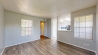 This single-floor, 1-bedroom, 1-bathroom unit is thoughtfully designed to maximize its compact footprint. The entry hallway, finished with wood-style flooring, provides a welcoming transition into the open living area. The living room is bright and airy, featuring multiple windows with horizontal blinds that allow ample natural light to fill the space. The wood-style flooring continues throughout, complemented by neutral gray walls and a textured ceiling. A half-wall pass-through connects the living room to the kitchen, which is a standout feature of the unit. The kitchen is outfitted with white cabinetry, white countertops, stainless steel appliances including a dishwasher, range, and microwave, and a striking blue mosaic tile backsplash that adds a pop of color. The bedroom is generously sized and features plush carpet flooring, a window for natural light, and direct access to a walk-in closet with built-in shelving and hanging rods. The bathroom includes a full tub-shower combo with white tile surround and clean, simple fixtures. Overall, the unit presents a clean, modern aesthetic throughout with consistent finishes and a practical, well-organized layout.
