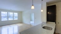 This single-floor space offers an open, airy layout that seamlessly connects the kitchen and living areas. The kitchen is anchored by a granite countertop with a stainless steel sink, complemented by pendant lights that hang gracefully overhead, providing both functional and ambient lighting. The light-toned hardwood flooring extends throughout the main living area, reflecting natural light and enhancing the sense of spaciousness. A window along the far wall invites abundant natural light, brightening the neutral, cream-toned walls. The open passageway visible in the background connects to an additional room, suggesting a functional and well-organized floor plan. Vaulted or elevated ceilings add a sense of volume to the space, contributing to its open and welcoming atmosphere. The clean, minimal design aesthetic makes the space highly versatile and move-in ready.