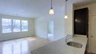 This single-floor space offers an open, airy layout that seamlessly connects the kitchen and living areas. The kitchen is anchored by a granite countertop with a stainless steel sink, complemented by pendant lights that hang gracefully overhead, providing both functional and ambient lighting. The light-toned hardwood flooring extends throughout the main living area, reflecting natural light and enhancing the sense of spaciousness. A window along the far wall invites abundant natural light, brightening the neutral, cream-toned walls. The open passageway visible in the background connects to an additional room, suggesting a functional and well-organized floor plan. Vaulted or elevated ceilings add a sense of volume to the space, contributing to its open and welcoming atmosphere. The clean, minimal design aesthetic makes the space highly versatile and move-in ready.