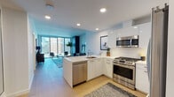 Located at 2801 Sunset Place in Los Angeles, this single-floor residence showcases a thoughtfully designed open-concept layout with a clean, contemporary aesthetic. Light wood flooring flows throughout the main living areas, complementing the crisp white walls and modern finishes that define the space.

The kitchen is a standout feature, outfitted with high-gloss white cabinetry extending to the ceiling, stainless steel appliances including a gas range, built-in microwave, and French door refrigerator, and a large island with a waterfall-edge white countertop that accommodates bar seating. Recessed lighting ensures the space is well-illuminated throughout.

The open living area benefits from expansive floor-to-ceiling windows that flood the room with natural light while offering elevated views of the surrounding Los Angeles cityscape. A teal accent wall adds a bold design touch, and the generous layout comfortably accommodates both a seating and lounge area.

The bedrooms continue the modern theme, each featuring large windows with city views and access to walk-in closet space. One bedroom is enhanced by a deep blue accent wall, adding warmth and character. The bathroom features large-format tile flooring, a wall-mounted toilet, and a clean, minimalist design. Overall, the unit presents a sophisticated urban living environment with polished, contemporary details throughout.